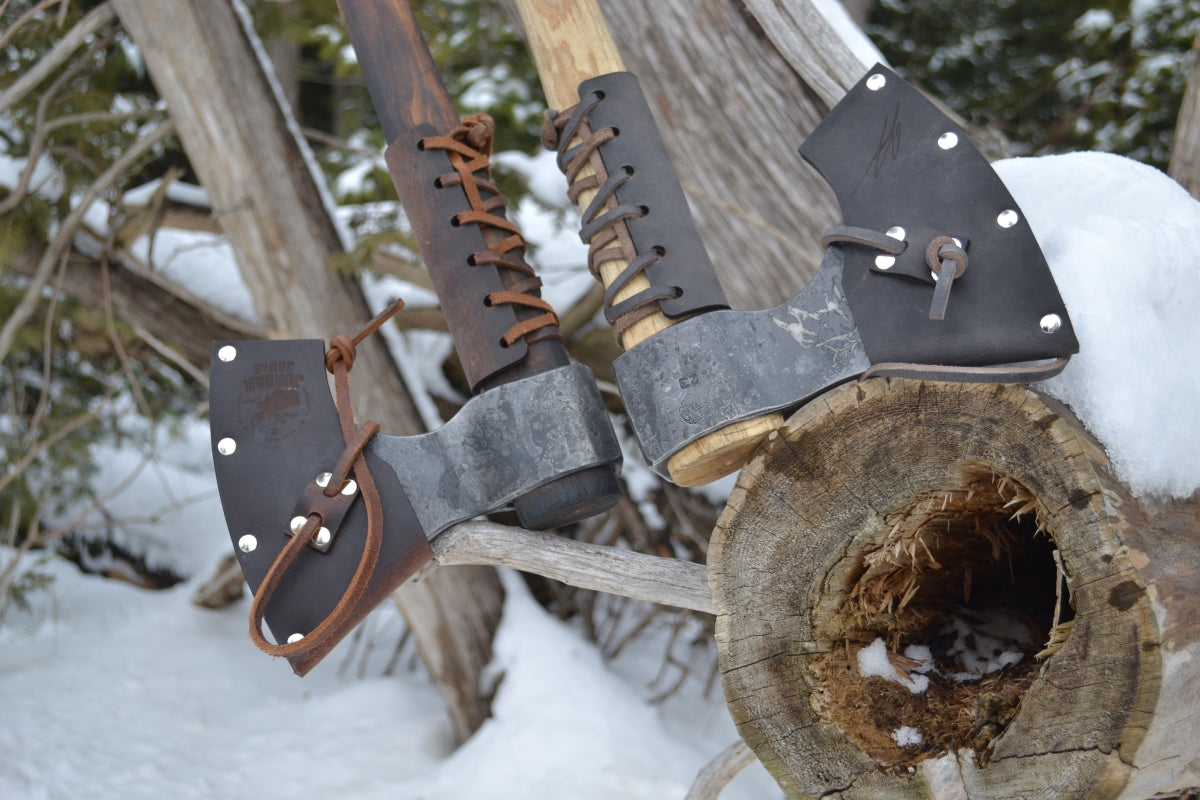 Taiga Moose axes with leather sheaths hanging on a wooden post in a snowy outdoor setting.