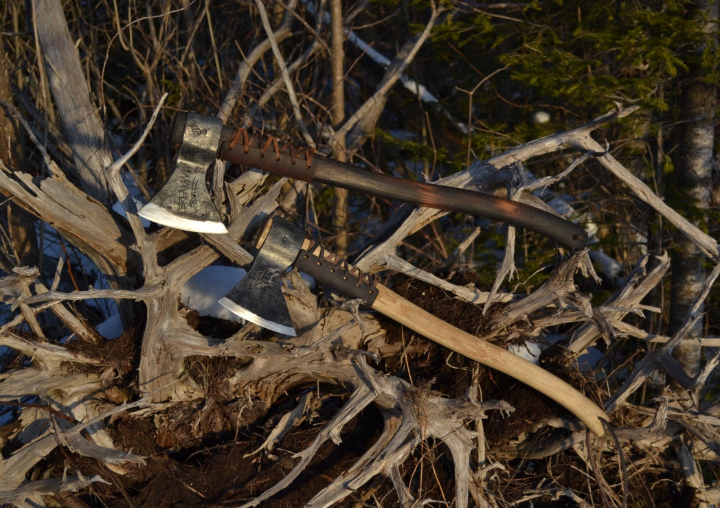 Two Taiga survival axes hanging on fallen tree root showing natural hickory and flame darkened handle finish options by Broad River Forge